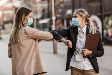 Businesswomen with safety masks greeting with elbow bump in front of office building.