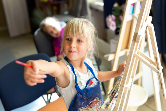 Cute blond smiling caucasian kid painting on wooden easel in class workshop lesson at art studio. Little girl holding brush in hand and having fun drawing with paints. Child early development concept