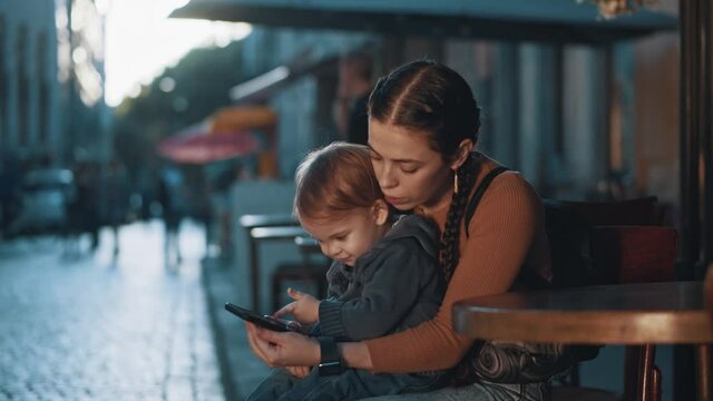 Happy young mom with little son watching fun videos on smartphone playing games on digital devide together. Outdoor walk. Motherhood. Kids.