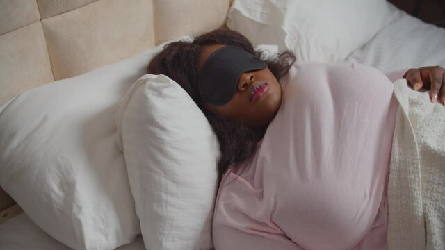 Beautiful plump african american woman wearing sleep eye mask, lying on uncomfortable pillows and hard mattress, moving from side to side, feeling restlessness and discomfort in bed during sleeping.