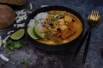 Vegan thai red curry  sauce with tofu, radddish and carrot over rice in black bowl with fork and spoon