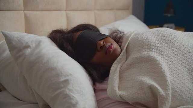 Restless charming chubby black woman wearing sleeping mask, lying on bed suffering from sleep disorder and insomnia, throwing away uncomfortable pillow while resting at night in bedroom.