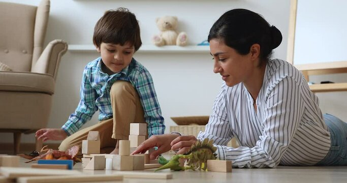 Indian nanny spend time in modern cozy home with little boy. Loving caring mother play with preschooler son with dinosaurs, construct tower use wooden bricks set. Child development, playtime concept