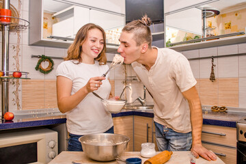 Happy young family, husband and wife, prepare cookies in light European kitchen, wife feeds her husband with raw dough