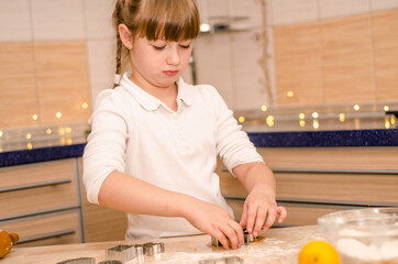 Cute happy girl learns how to cook Christmas gingerbread cookies in bright cozy kitchen, cuts cookies with molds