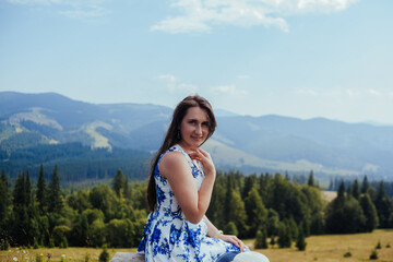 Pretty woman in elegant white and blue dress sitting on bench in the mountains and smiling. Beautiful girl relaxing in the top of Carpathians. travel to the mountains on a warm sunny summer's day