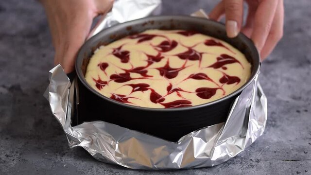 Woman preparing cheesecake with berry sauce in the kitchen. Wrapping the aluminum foil around the springform pan.