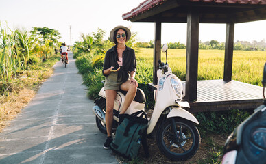 Fototapeta na wymiar Portrait of cheerful female tourist in sunglasses and straw hat smiling at camera while resting on vintage moped parked near rice fields and gazebo, millennial hipster girl with cellphone laughing