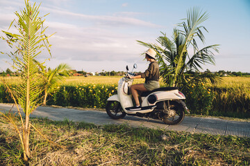 Side view of casual dressed female tourist drive motorcycle exploring Indonesia during summertime for getaway travel trip, young hipster girl getting to destination on Bali via vintage rent scooter © BullRun