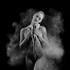 A beautiful slender ballet dancer girl wearing a bodysuit, sensually holds pointe shoes in her hands among the flying flour which covers her body, on black. Artistic, commercial, monochrome design