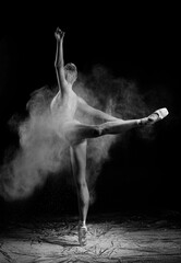 A beautiful slender ballet dancer girl wearing a bodysuit and pointe shoes, posing dancing among the clouds of flying flour on a black background. Artistic, commercial, monochrome design