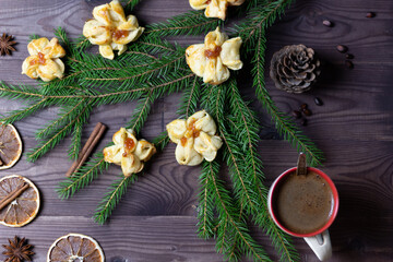 cookies of flower shape on christmas fir tree branches with cinnamon, star anis, dry orange slices, cup of coffee, pine cone, nuts, jam on brown wooden background