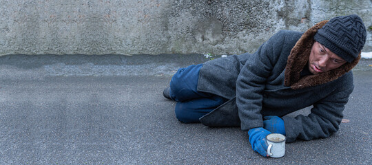 A homeless man lying on the asphalt reaches for a mug of tea against the background of a gray wall.