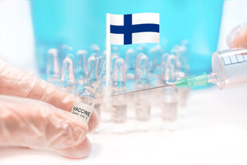 Hands holding a vaccine vial and a syringe with rows of the same capsules and a flag of Finland in the background, illustrating plans for global vaccination against Covid-19 (SARS-CoV-2, coronavirus).