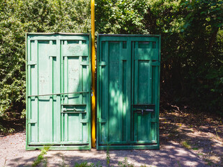 Two green iron cargo containers standing on the street with a yellow chimney passing between them against the backdrop of green trees