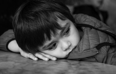 Black and white photo of Kid with bored face,Lonely boy putting his chin down on table and looking...