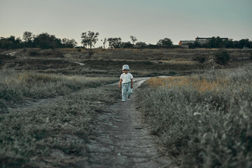 Obraz premium Two-year-old child in a hat walks along a country road, summer. Knowledge of the world.