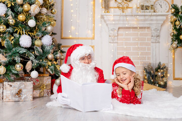 Santa Claus reads a book to a child girl at the Christmas tree, the concept of new year and Christmas