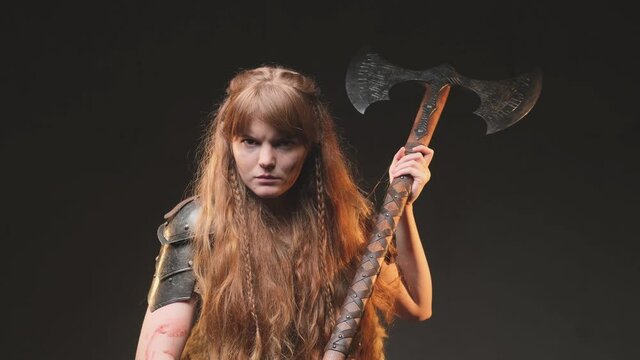 Historical portrait of a savage and angry female barbarian from nord holding a two handed double axe in dark background. Military scandinavian woman with long brown hairs dressed in antique steel