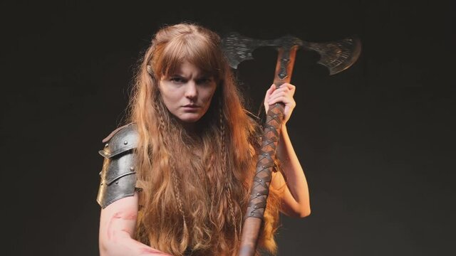 Historical portrait of a savage and angry female barbarian from nord holding a two handed double axe in dark background. Military scandinavian woman with long brown hairs dressed in antique steel