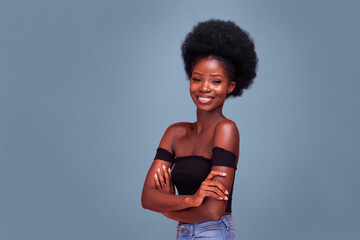 Smiling Confident young African American girl with afro hair standing with arms folded in black bare shoulder top and blue denim jeans. Isolated on dirty blue background.