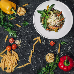 Vegetable pasta sprinkled with parmesan and garnished with basel. Nearby are sprinkled pasta, of which an arrow is laid out to a plate. Nearby are Bulgarian peppers, zucchini and tomatoes, greens.