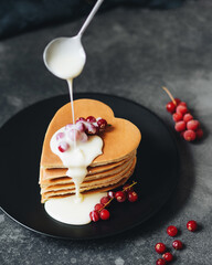 Breakfast for lovers. Pancakes in the form of a heart. Pour sweet cream on top with a spoon and garnish with fresh red currant berries. Ideas for Valentine's Day. American pancakes are stacked.