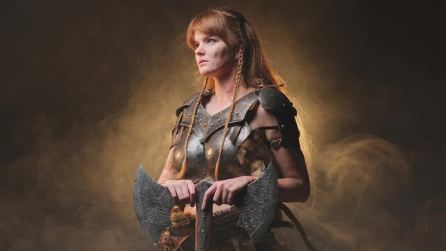 Beautiful and at the same time dangerous female viking warrior dressed in steel light armour holding a two handed double axe in dark atmospheric background with fog. Warlike and antique portrait of a