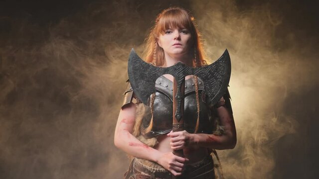 Beautiful and at the same time dangerous female viking warrior dressed in steel light armour holding a two handed double axe in dark atmospheric background with fog. Warlike and antique portrait of a