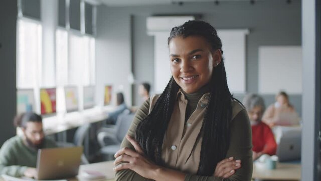 Waist up portrait shot of young cheerful Afro-American businesswoman with braided hair standing with arms crossed in office, looking at camera and smiling