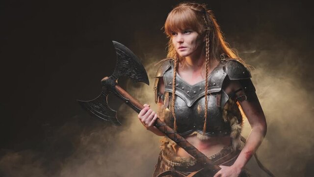 Martial and violent female viking barbarian armed with steel double axe and with long brown hairs posing in dark background with fog. Ready to fight nordic valkyrie armed with huge axe.