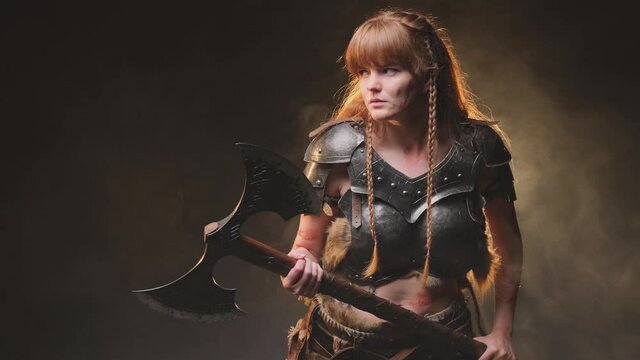 Martial and violent female viking barbarian armed with steel double axe and with long brown hairs posing in dark background with fog. Ready to fight nordic valkyrie armed with huge axe.