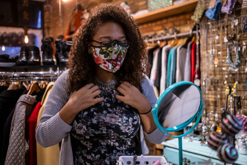 African American woman wearing a mask is inside a store trying on a new necklace she wish to purchase. she wears glasses with and has long curly hair to make herself beautiful during covid-19