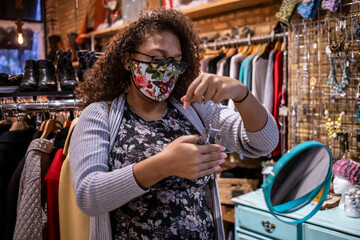 African American woman wearing a mask is inside a store trying on a new necklace she wish to purchase. she wears glasses with and has long curly hair to make herself beautiful during covid-19