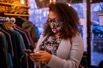 African American mixed woman with curly hair and wearing a protective mask is texting on her smart phone while shopping during Covid-19 pandemic. 