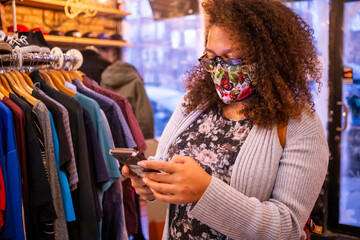 African American mixed woman with curly hair and wearing a protective mask is texting on her smart phone while shopping during Covid-19 pandemic. 