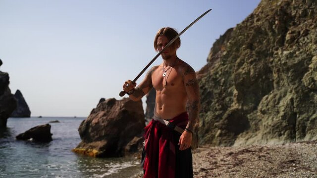 Mature Tattoed Man Holding Swords Ready To Fight on the rocky sea background - Portrait Of A Handsome Muscular Ancient Warrior With A Swords. Martial art concept