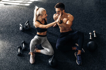 young man and woman sitting on the floor and giving each other five in the gym, fitness people after workout in the gym