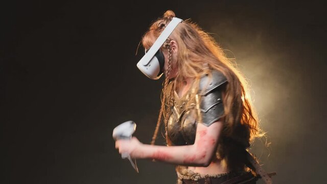 Female gamer with long brown hairs and dressed in antique steel armour weared with virtual reality glasses poses in dark background playing and imaging battle holding joysticks in dark and smokey