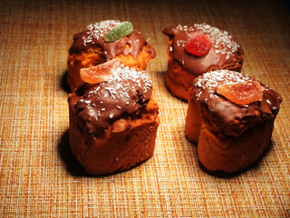 Four homemade cupcakes lie on the tablecloth. Cupcakes are decorated with chocolate icing, coconut chips, and pieces of marmalade.