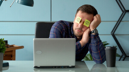 Tired comic male manager pretends working sleeping with stickers on face sits at desk with laptop. 