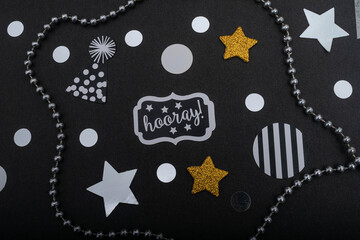 Silver necklace, golden and silver stars, little hats and confetti on black background. New Year and party decoration