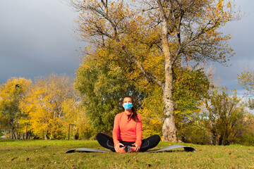 Naklejka na ściany i meble Sporty dressed woman doing stretching in the lotus flower position on a mat in the park wearing a mask to protect herself from the coronavirus