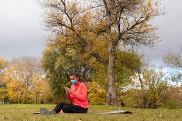 Woman dressed in sport sitting on a mat in the park wearing a mask to protect herself from the coronavirus makes a video call or checks her mobile