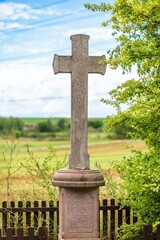 Fototapeta na wymiar Zadni Kopanina, Czech Republic - August 23 2020: A stone cross standing inside a wooden fence in field with green tree. Colorful background. Sunny summer day with blue sky and clouds. Vertical image.