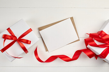 Greeting card mockup with paper envelope and red ribbon and gift box on white table