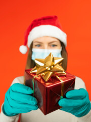 Young woman with protective mask and protective gloves holds a Christmas present on red Christmas background, look straight ahead Christmas with social distancing