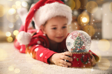 Little girl in Santa hat playing with snow globe on floor. Bokeh effect