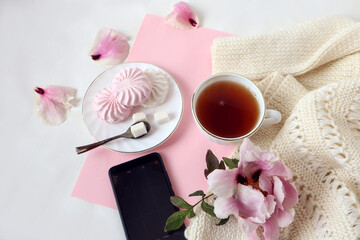 A Cup of tea on a white knitted sweater with a delicate peony flower, soft pink marshmallows, phone, top view-the concept of pleasant phone greetings