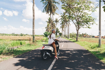 Stylish young woman driving motorbike on road in tropical country
